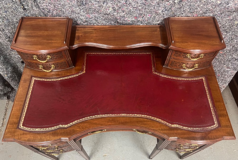 A mahogany writing desk featuring a red leather inset surface, carved drawer fronts, brass swanneck handles, and an elevated superstructure with twin drawer units. View VI