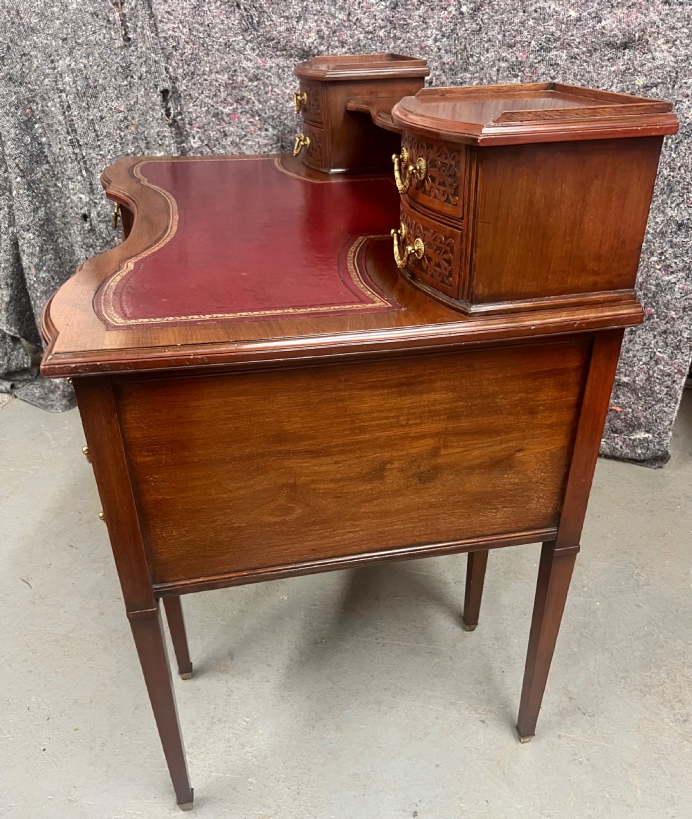 A mahogany writing desk featuring a red leather inset surface, carved drawer fronts, brass swanneck handles, and an elevated superstructure with twin drawer units. View VIII