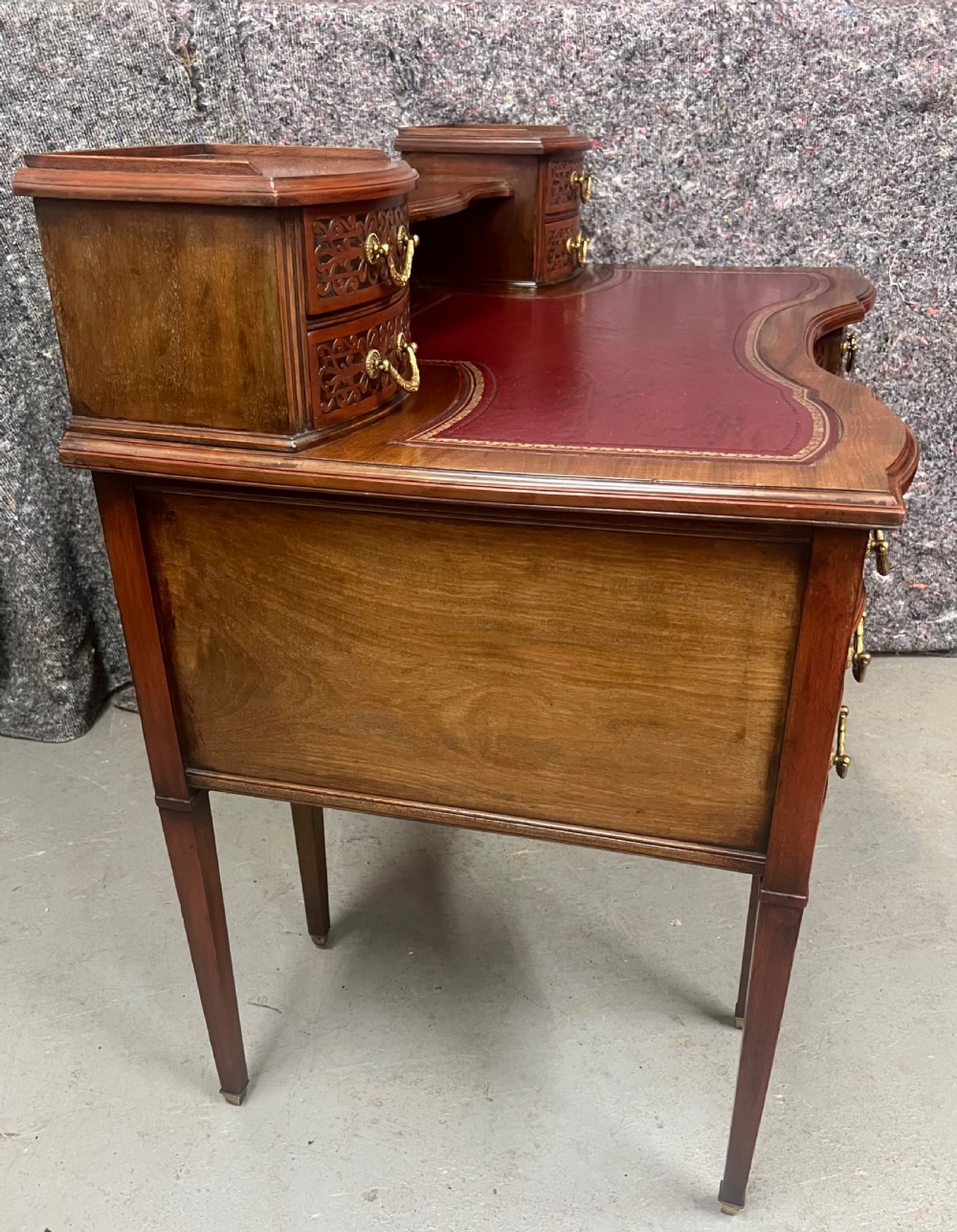 A mahogany writing desk featuring a red leather inset surface, carved drawer fronts, brass swanneck handles, and an elevated superstructure with twin drawer units. View IX