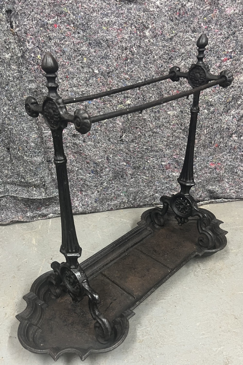A black Victorian cast iron stick stand featuring twin horizontal support bars, urn finials, rosette mouldings, and a scalloped drip tray base.