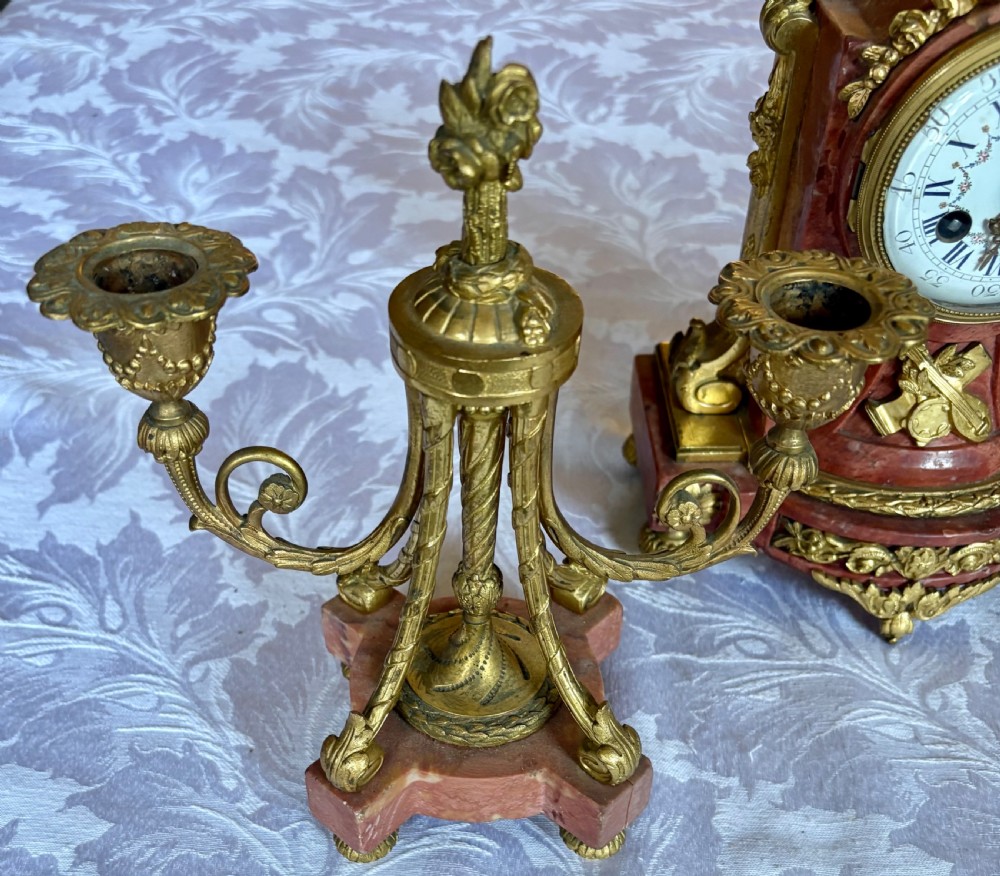 A three-piece French clock garniture featuring a central pink marble mantel clock decorated with gilt bronze floral mounts and flanked by two matching twin-light candelabra. View IX