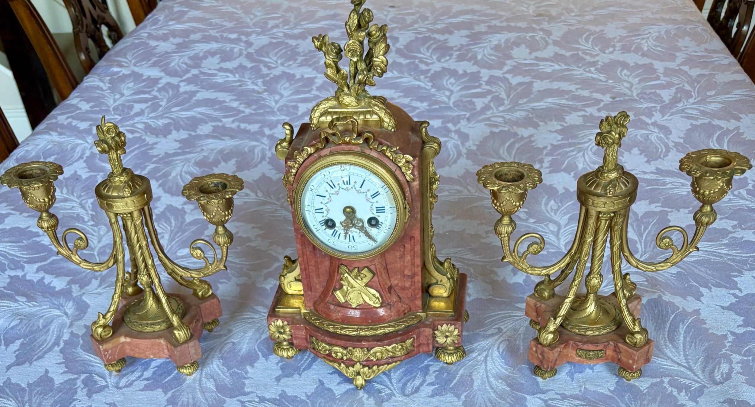 A three-piece French clock garniture featuring a central pink marble mantel clock decorated with gilt bronze floral mounts and flanked by two matching twin-light candelabra.