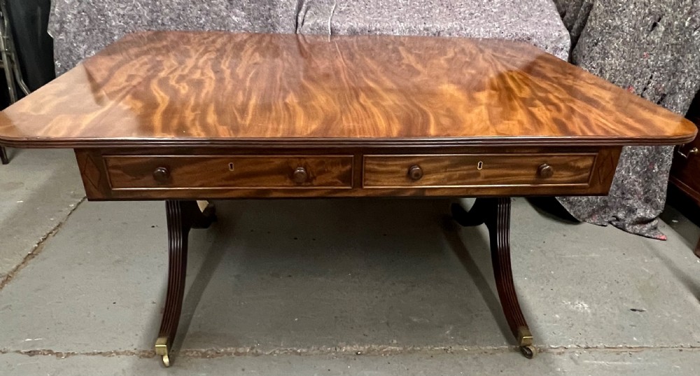 A mahogany library table featuring a flame veneer surface, frieze drawers with turned knobs, and reeded sabre legs resting on brass castors.