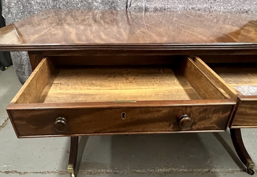 A mahogany library table featuring a flame veneer surface, frieze drawers with turned knobs, and reeded sabre legs resting on brass castors. View X