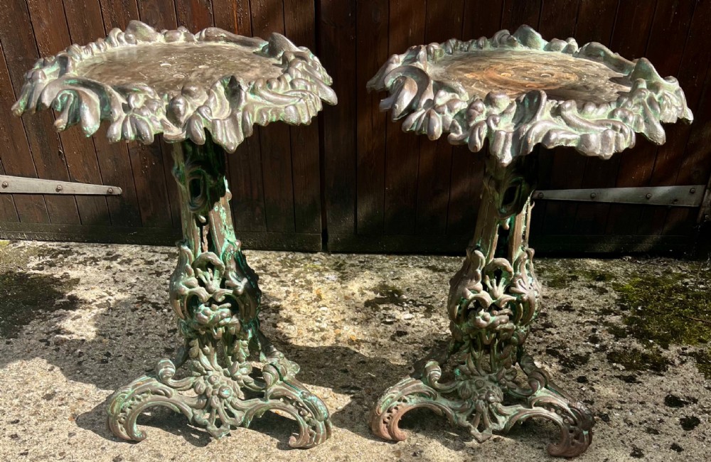 A pair of ornate cast iron pedestal tables with rococo scrollwork, tripod bases, and a verdigris green patinated finish.