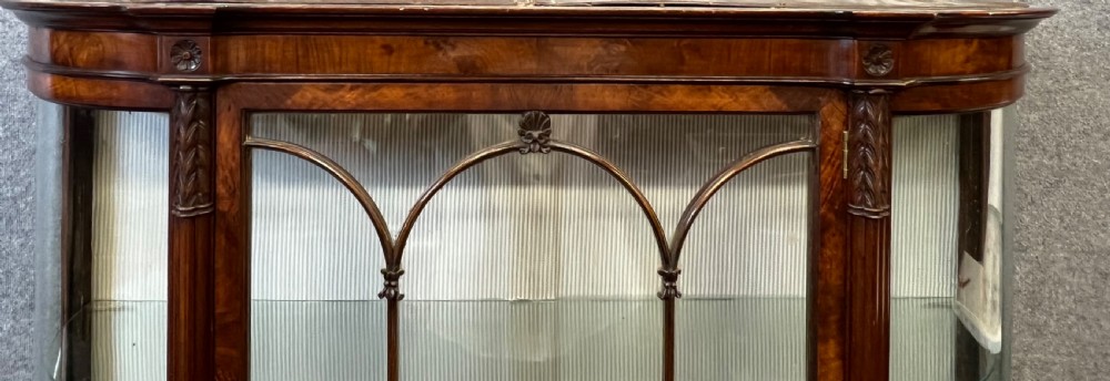 An Edwardian burr walnut display cabinet featuring curved glass side panels, an astragal glazed central door, glass shelves, and cabriole legs. View X