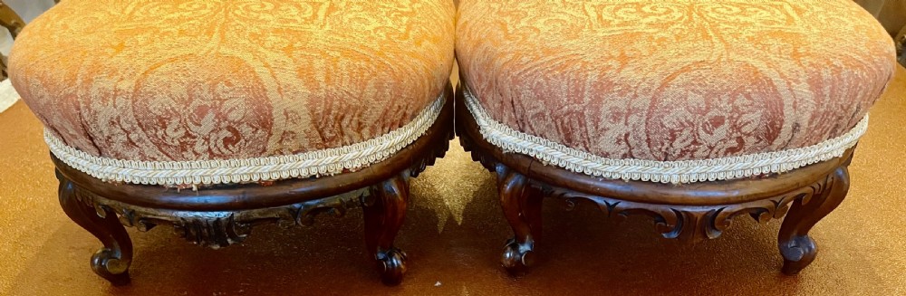 A matching pair of low circular Victorian footstools with patterned orange upholstery, decorative braid trim, and dark carved wooden legs. View II