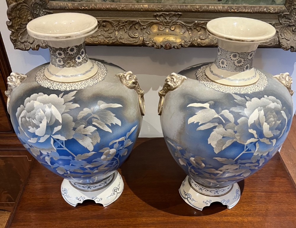 A pair of large Japanese Satsuma earthenware vases featuring painted blue and grey peonies, molded lion head handles, and a crackled cream glaze.
