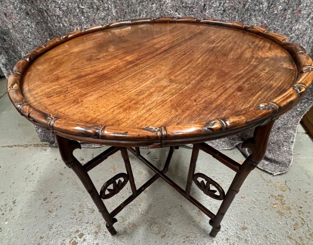 An oval brown hardwood coffee table with carved faux bamboo trim on a folding base.