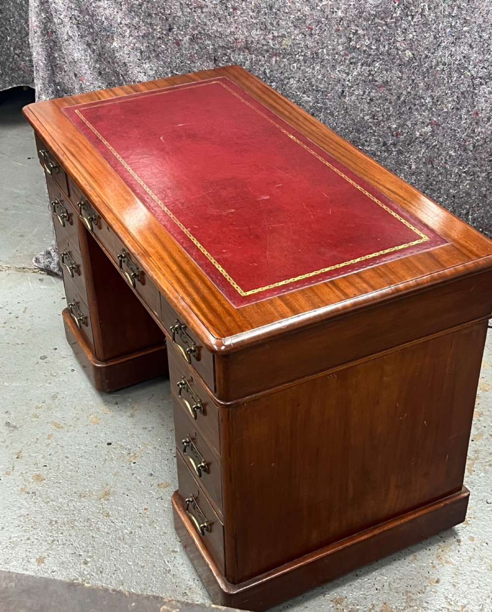 A compact Victorian mahogany pedestal desk featuring a red leather inset top, nine drawers with brass handles, and a plinth base on castors. View II