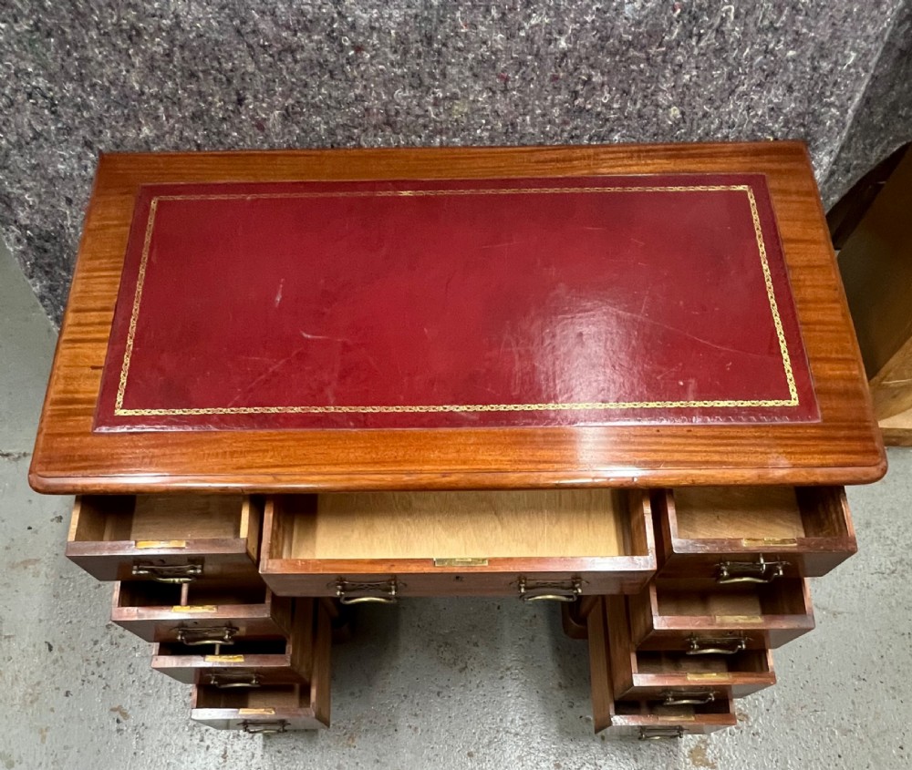 A compact Victorian mahogany pedestal desk featuring a red leather inset top, nine drawers with brass handles, and a plinth base on castors. View III