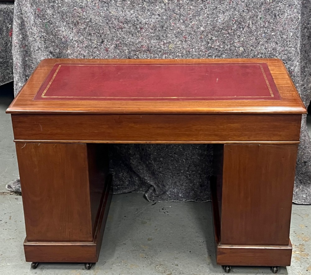 A compact Victorian mahogany pedestal desk featuring a red leather inset top, nine drawers with brass handles, and a plinth base on castors. View V