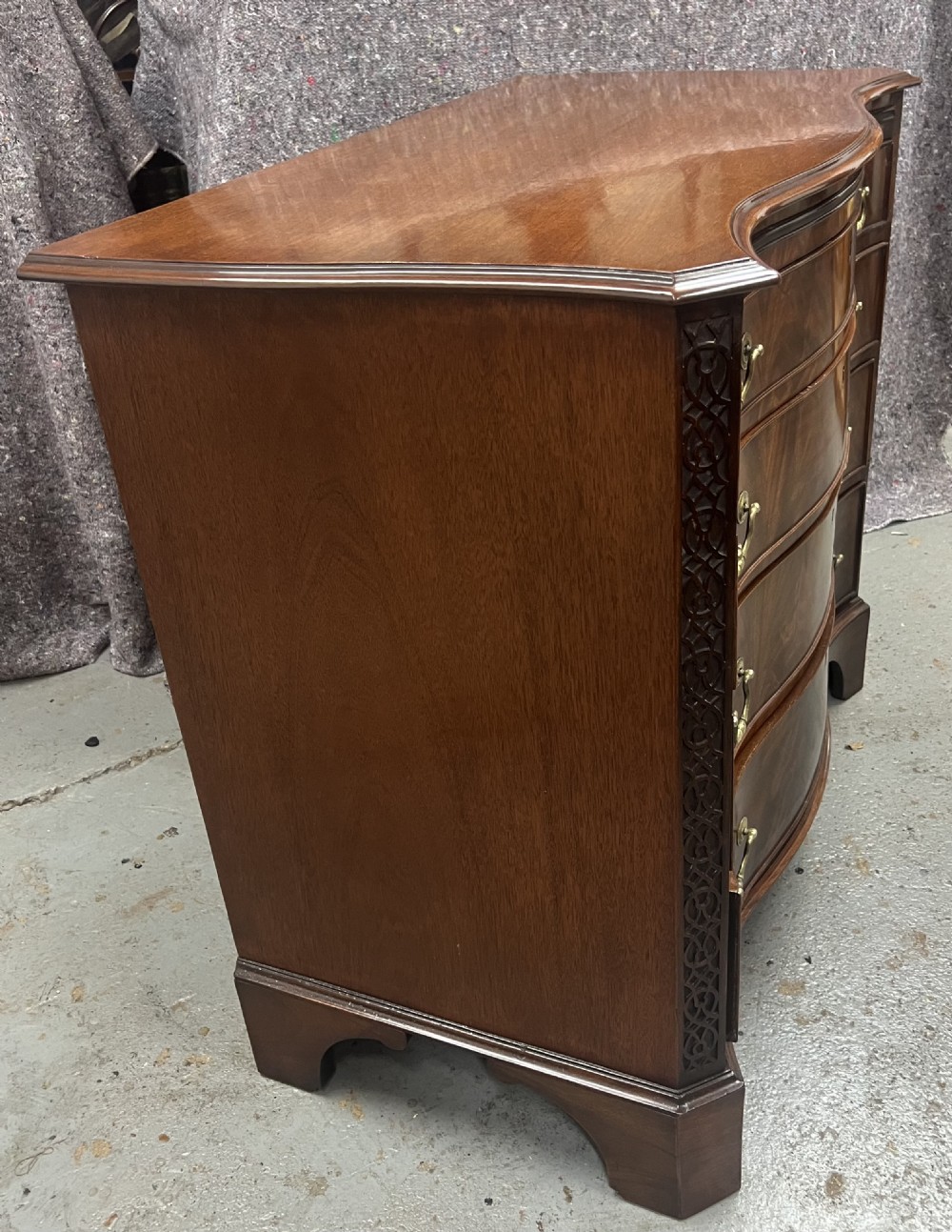 A Chippendale style mahogany chest of drawers featuring a serpentine front facade, carved blind fretwork canted corners, brass swan-neck handles, and bracket feet. View V
