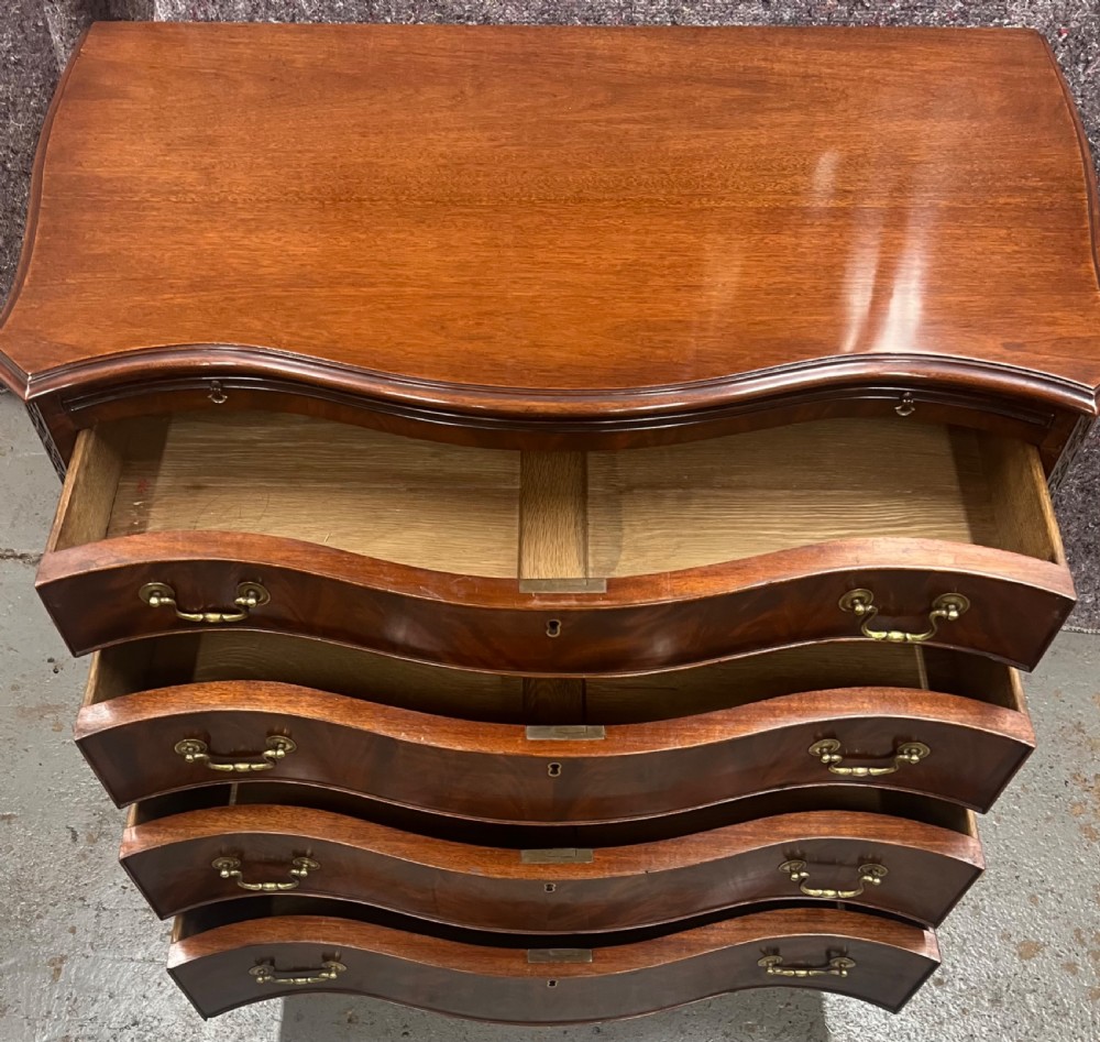 A Chippendale style mahogany chest of drawers featuring a serpentine front facade, carved blind fretwork canted corners, brass swan-neck handles, and bracket feet. View VI