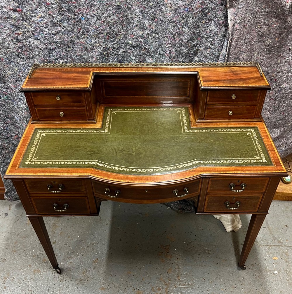 An Edwardian inlaid mahogany writing desk featuring a green leather inset writing surface, raised drawered superstructure, brass gallery top, and tapered square legs with castors.