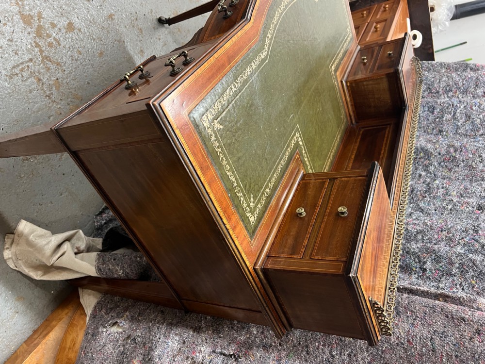 An Edwardian inlaid mahogany writing desk featuring a green leather inset writing surface, raised drawered superstructure, brass gallery top, and tapered square legs with castors. View V