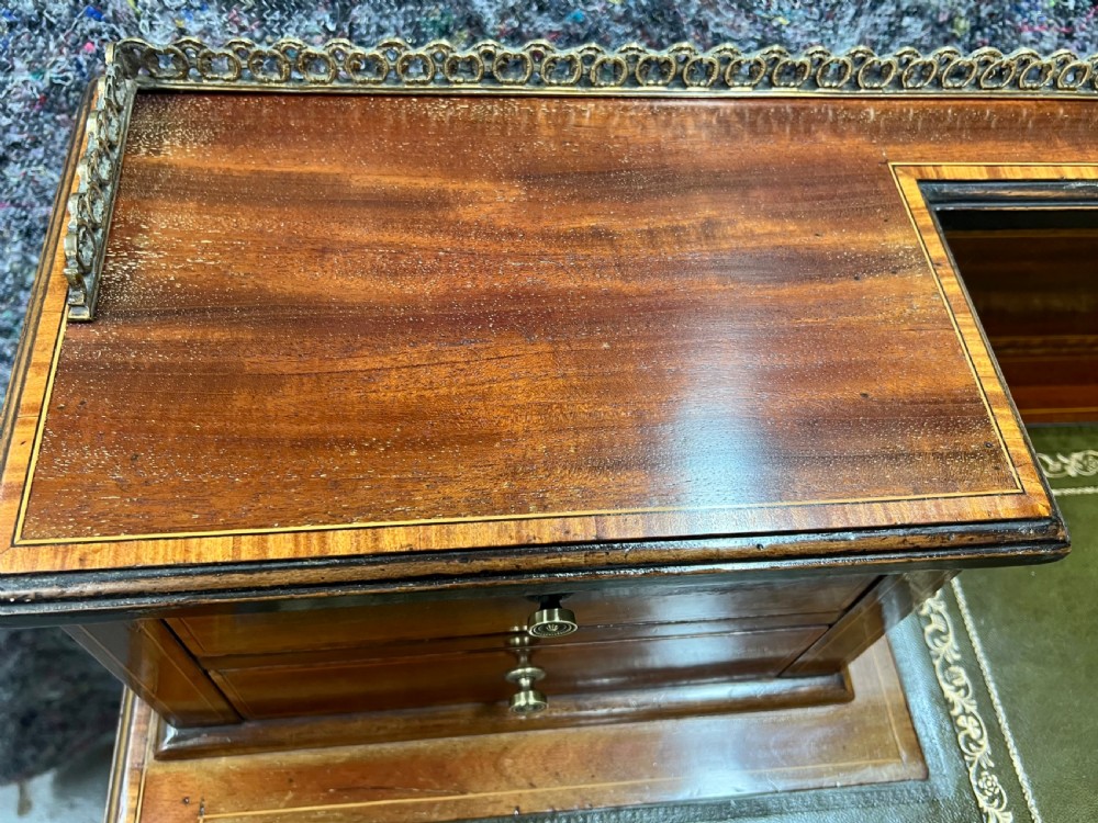 An Edwardian inlaid mahogany writing desk featuring a green leather inset writing surface, raised drawered superstructure, brass gallery top, and tapered square legs with castors. View VII