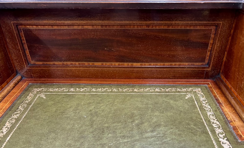 An Edwardian inlaid mahogany writing desk featuring a green leather inset writing surface, raised drawered superstructure, brass gallery top, and tapered square legs with castors. View VIII