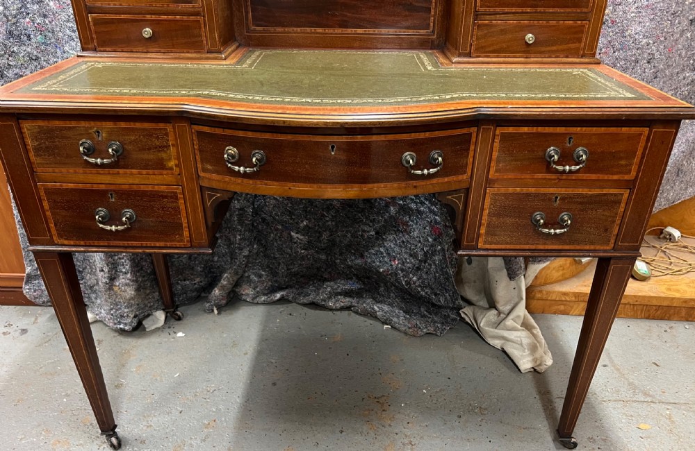 An Edwardian inlaid mahogany writing desk featuring a green leather inset writing surface, raised drawered superstructure, brass gallery top, and tapered square legs with castors. View IX