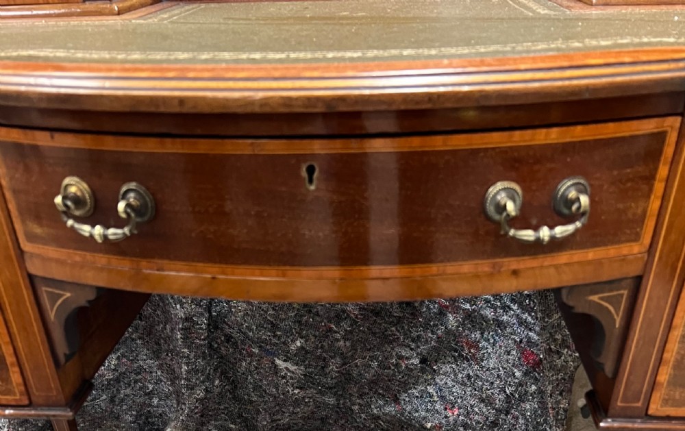 An Edwardian inlaid mahogany writing desk featuring a green leather inset writing surface, raised drawered superstructure, brass gallery top, and tapered square legs with castors. View X