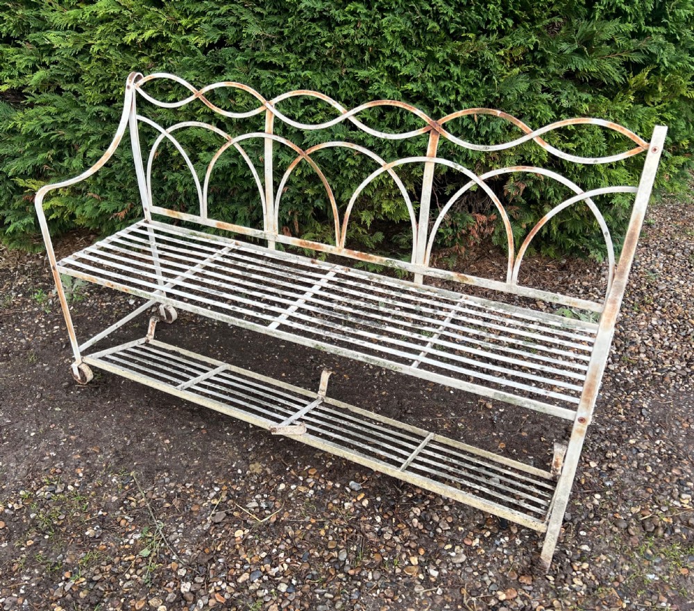 An off-white painted wrought iron garden seat featuring slatted strapwork construction, gothic backrest arches, and a lower footrest with weathered patina.