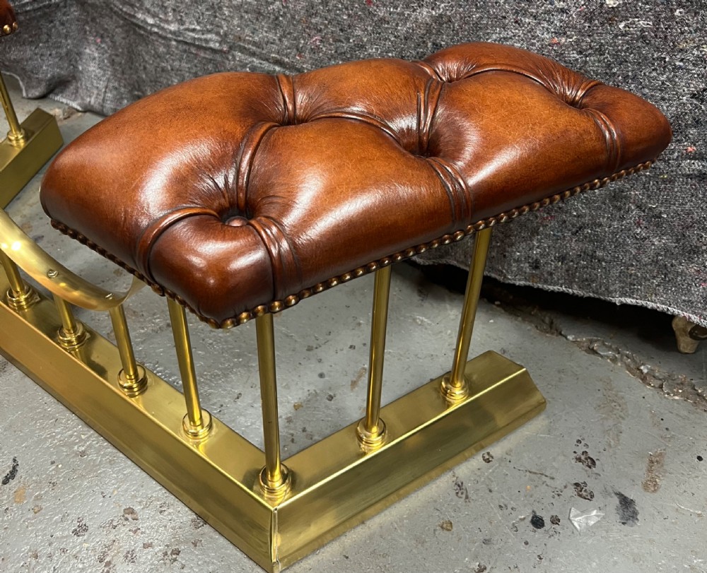 A brass fireplace club fender featuring two deep-buttoned brown leather seats supported by vertical brass spindles on a shaped metal base. View V