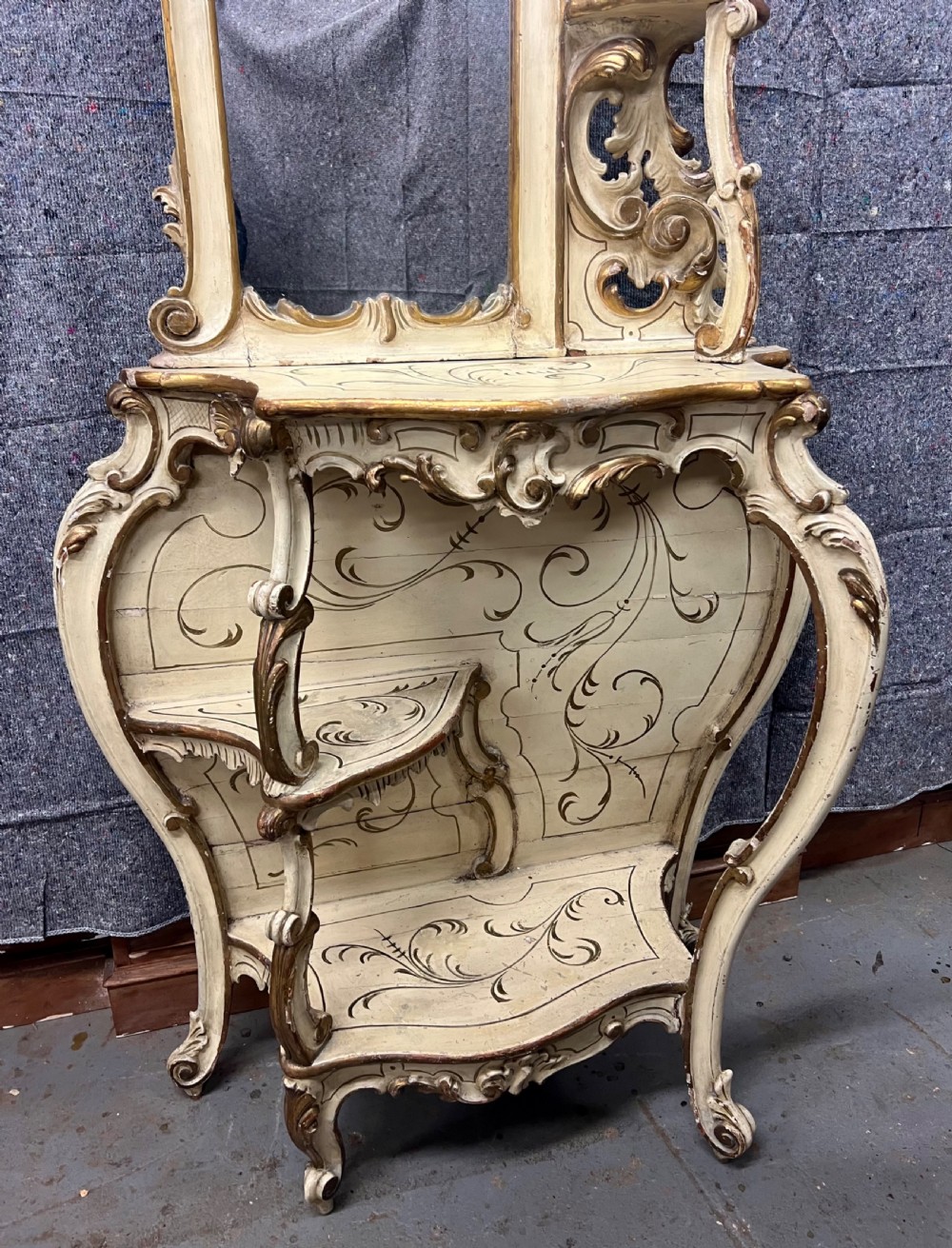 An ornate cream-painted and parcel-gilt Venetian Rococo console table featuring an asymmetrical tall mirror, tiered display shelves, and scrolling cabriole legs. View II