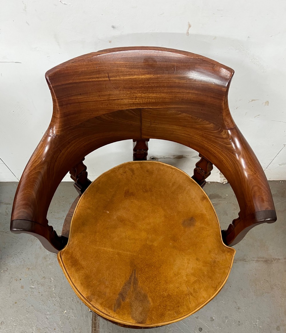 A Victorian mahogany revolving desk chair featuring a curved horseshoe backrest, carved support splats, tan suede upholstered seat, and cabriole-style legs on brass castors. View VI