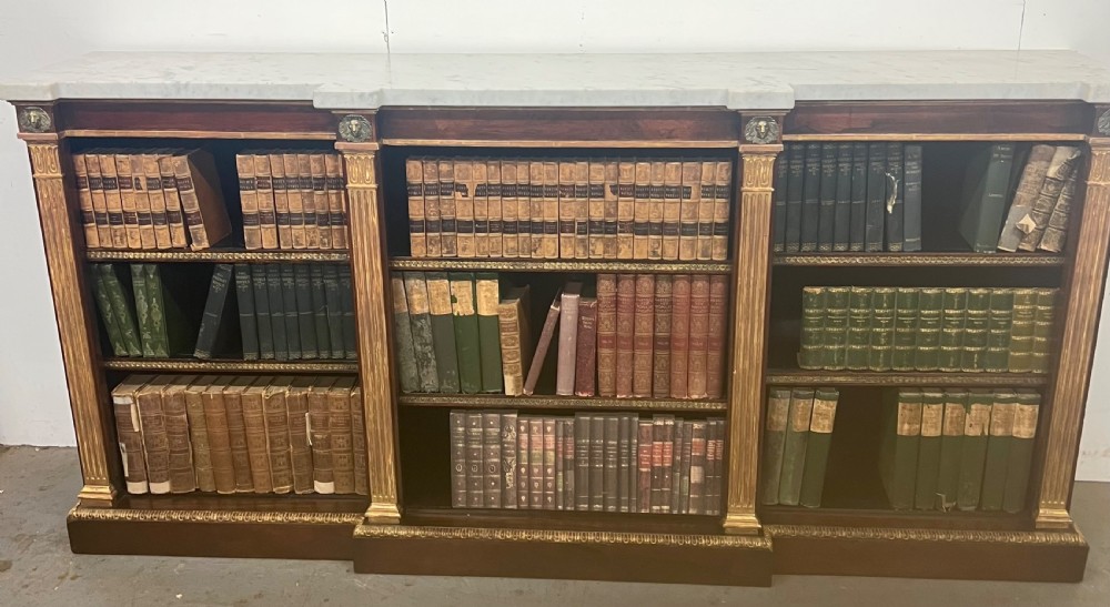 A low three-bay Regency style rosewood open bookcase with a white marble top, fluted gilded pilasters, and carved lion head mounts.