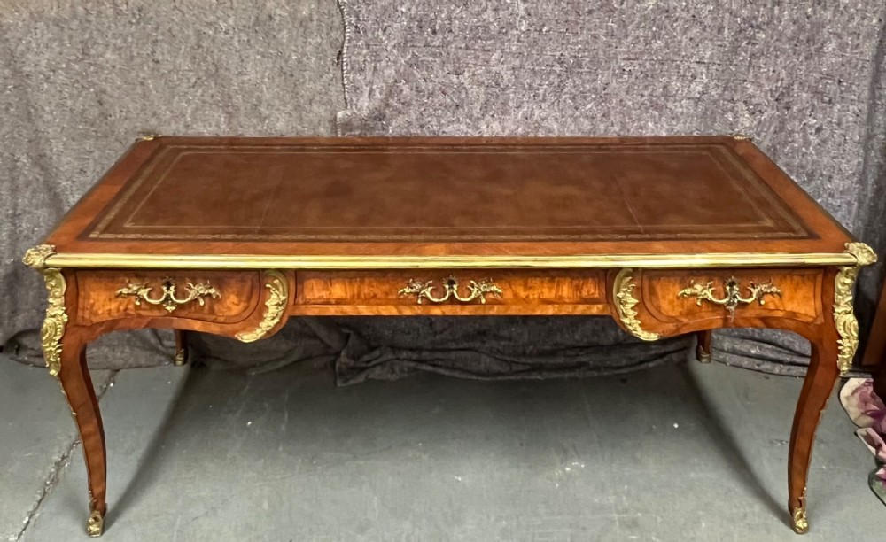 A nineteenth-century French bureau plat desk featuring wood marquetry, curved cabriole legs, ornate gilt-bronze mounts, three frieze drawers, and a leather inset writing top.