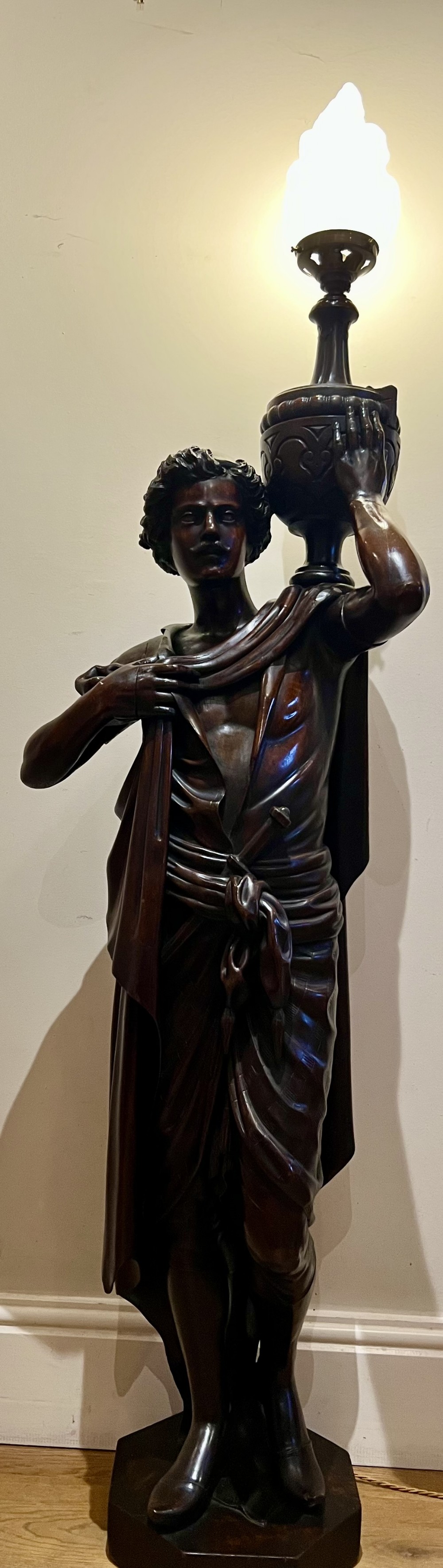 A 19th-century carved dark wood figural floor lamp depicting a draped male figure holding an illuminated flame glass urn on a octagonal base.