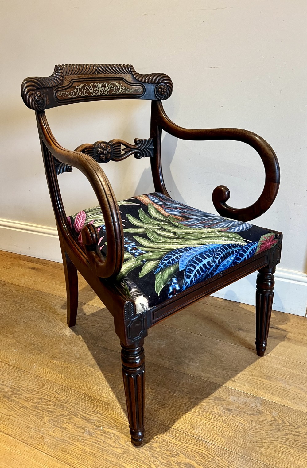 A Regency rosewood elbow chair featuring brass inlay along the top rail, curved scroll arms, a carved central splat, reeded front legs, and a colourful botanical upholstered seat.