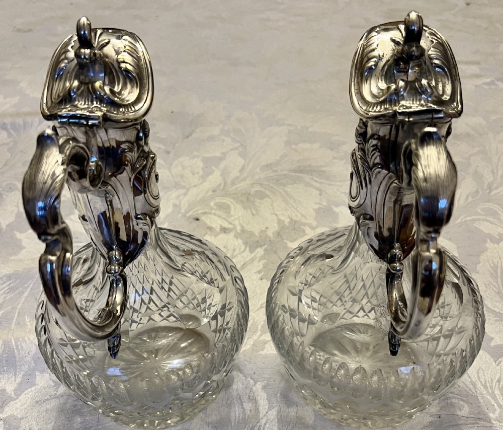 A pair of antique silver-plated claret jugs featuring ornate scroll handles, floral mounts, and diamond-pattern cut glass bodies. View III