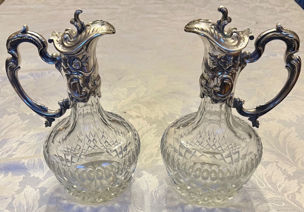 A pair of antique silver-plated claret jugs featuring ornate scroll handles, floral mounts, and diamond-pattern cut glass bodies. View IX