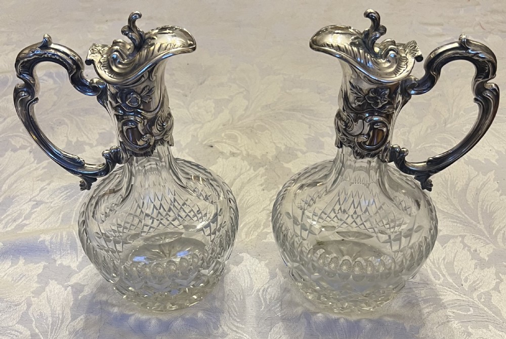 A pair of antique silver-plated claret jugs featuring ornate scroll handles, floral mounts, and diamond-pattern cut glass bodies. View X