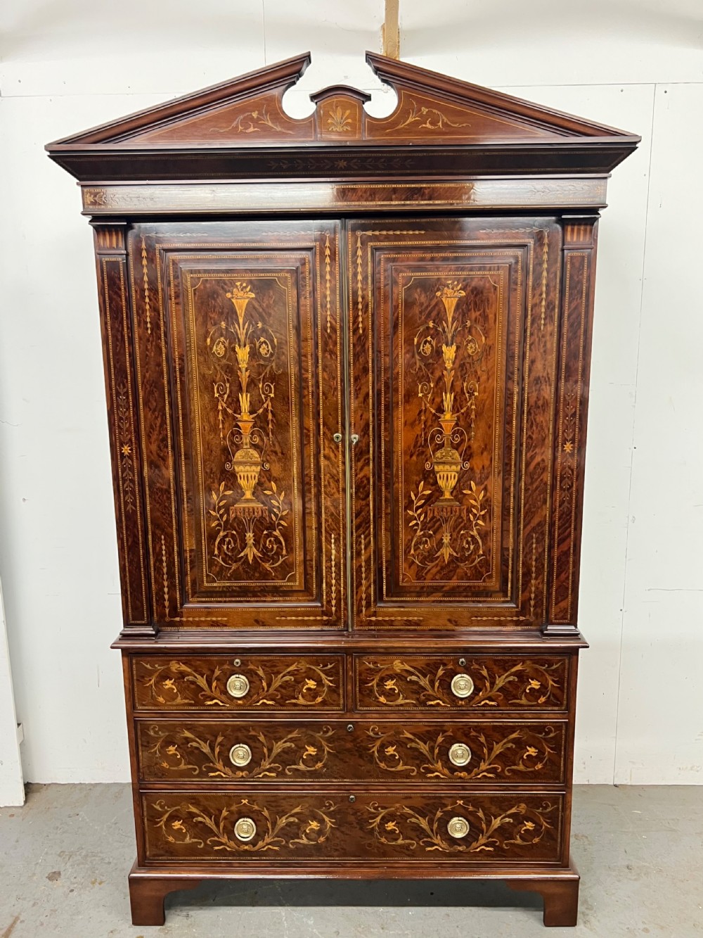 An antique dark mahogany linen press featuring classical marquetry urn inlay, a broken pediment top, two cabinet doors, and four lower drawers with circular brass handles.