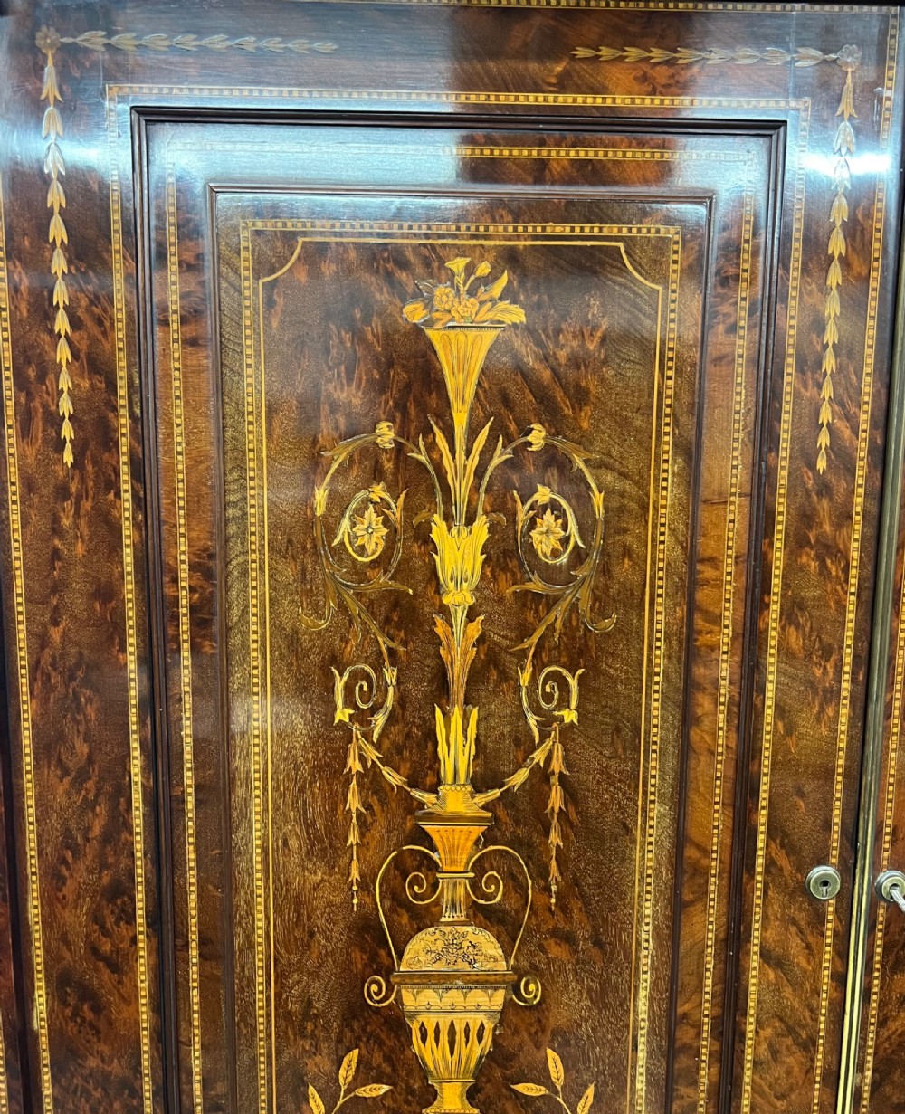 An antique dark mahogany linen press featuring classical marquetry urn inlay, a broken pediment top, two cabinet doors, and four lower drawers with circular brass handles. View IV