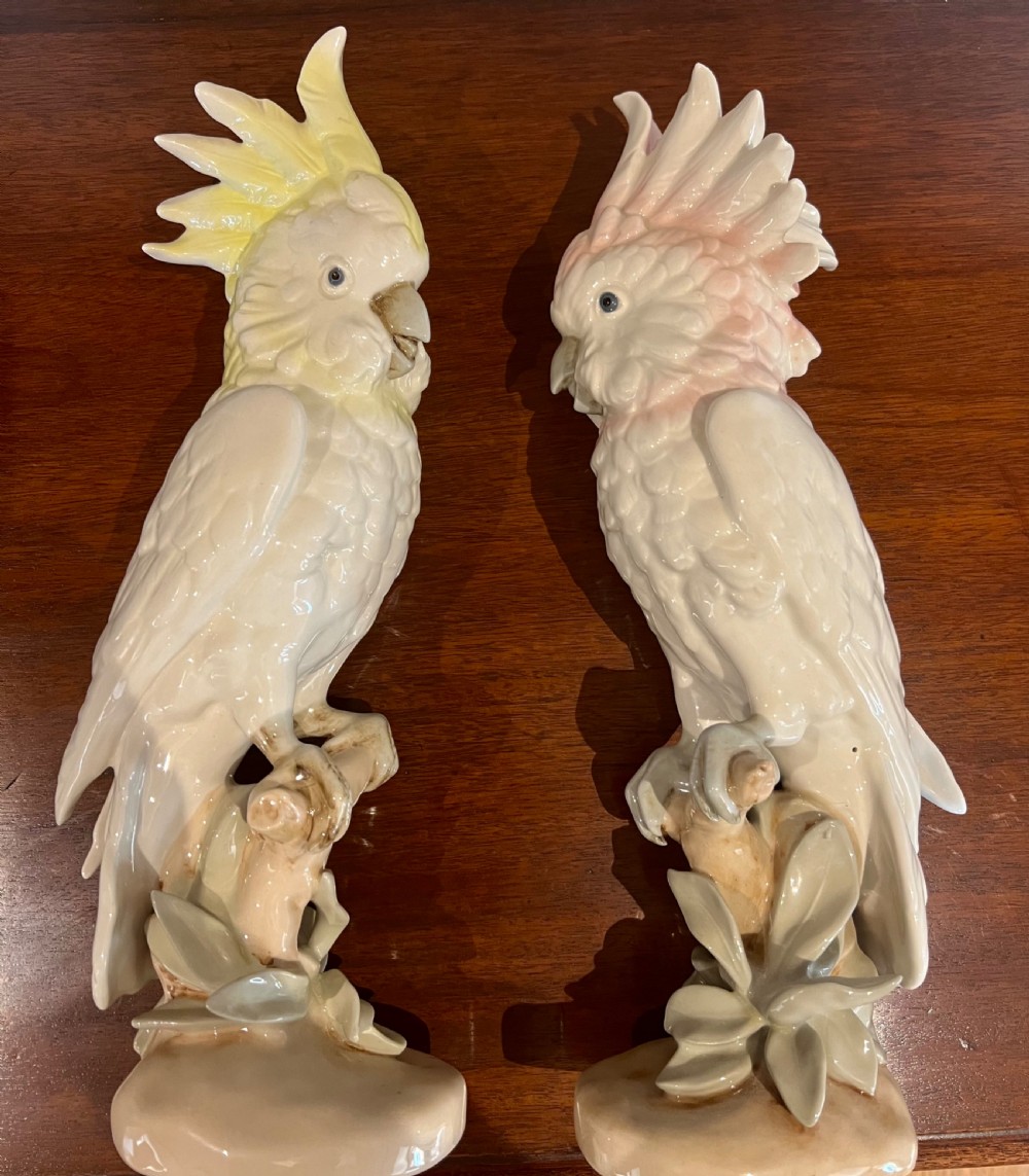 A pair of glazed porcelain cockatoo figurines perched on stylized foliage branches, one featuring a yellow crest and the other a soft pink crest.