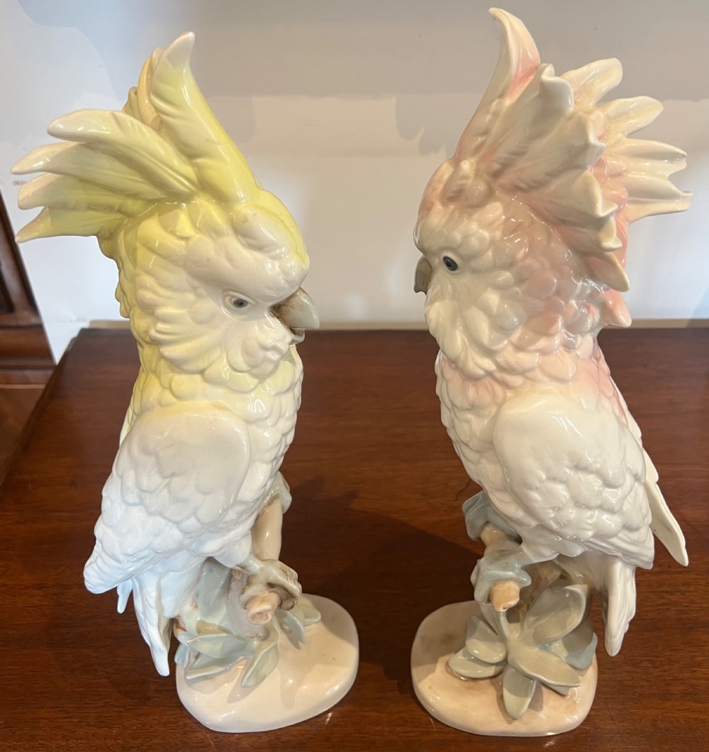 A pair of glazed porcelain cockatoo figurines perched on stylized foliage branches, one featuring a yellow crest and the other a soft pink crest. View X