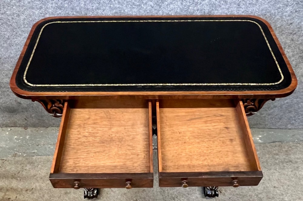 A Regency period dark rosewood freestanding writing table featuring a dark leather insert top, carved scroll accents, a turned central stretcher, and carved feet. View II