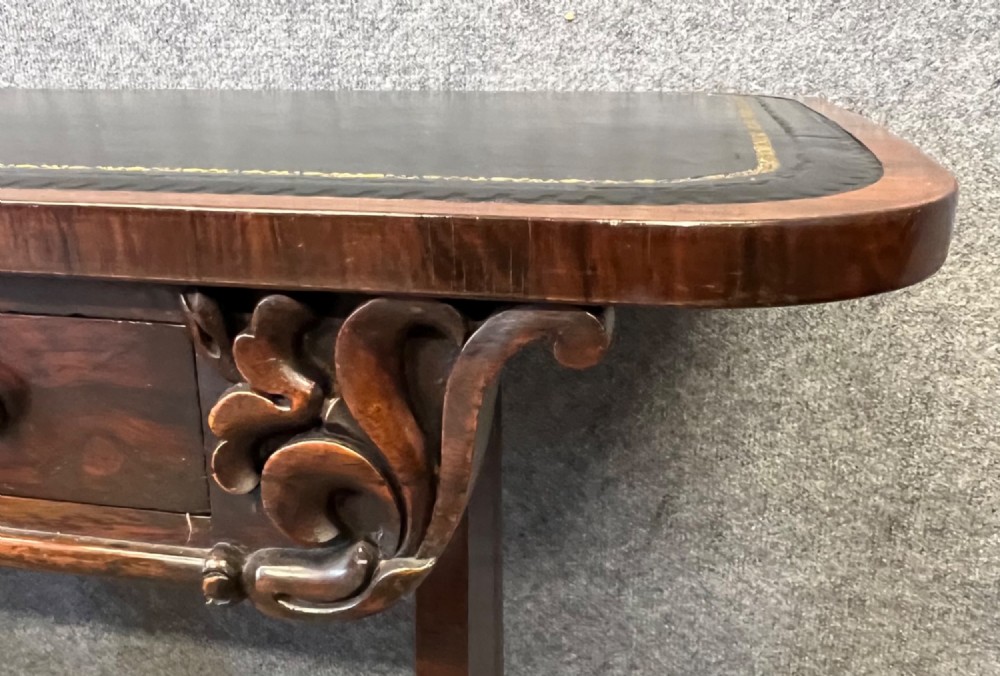 A Regency period dark rosewood freestanding writing table featuring a dark leather insert top, carved scroll accents, a turned central stretcher, and carved feet. View IV