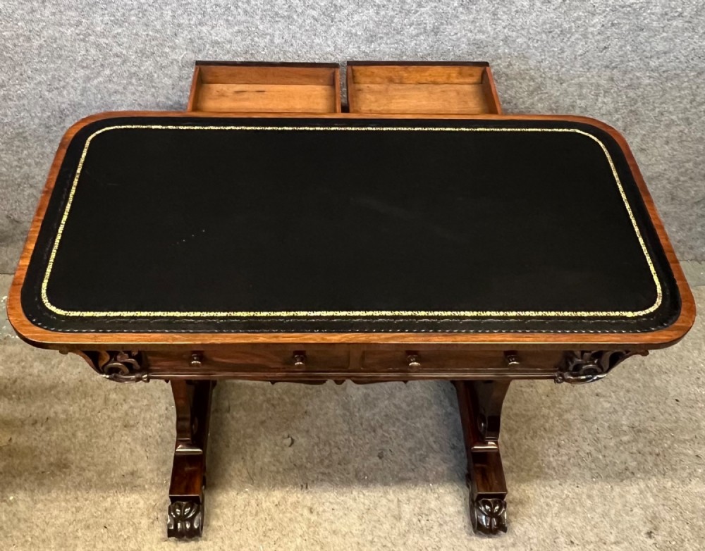 A Regency period dark rosewood freestanding writing table featuring a dark leather insert top, carved scroll accents, a turned central stretcher, and carved feet. View X