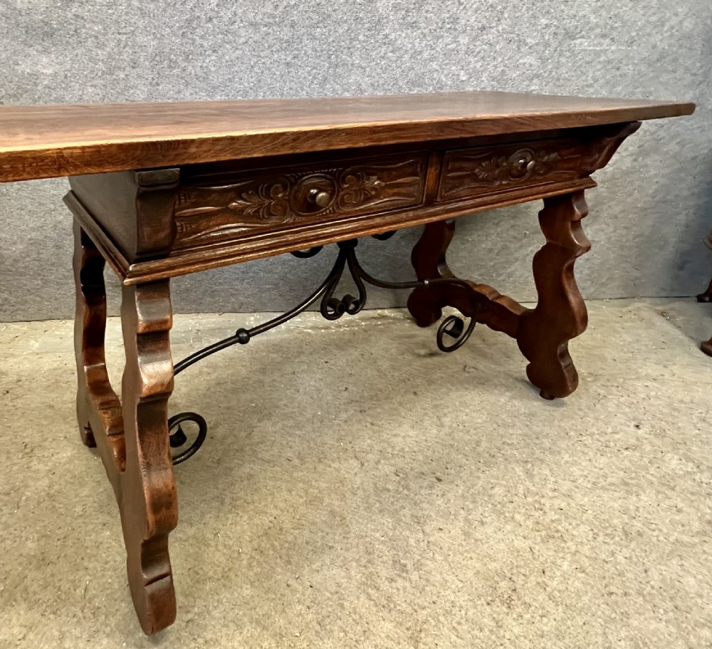 A dark oak Spanish console table featuring two carved drawers, lyre-shaped trestle legs, and a scrolled wrought iron stretcher. View X