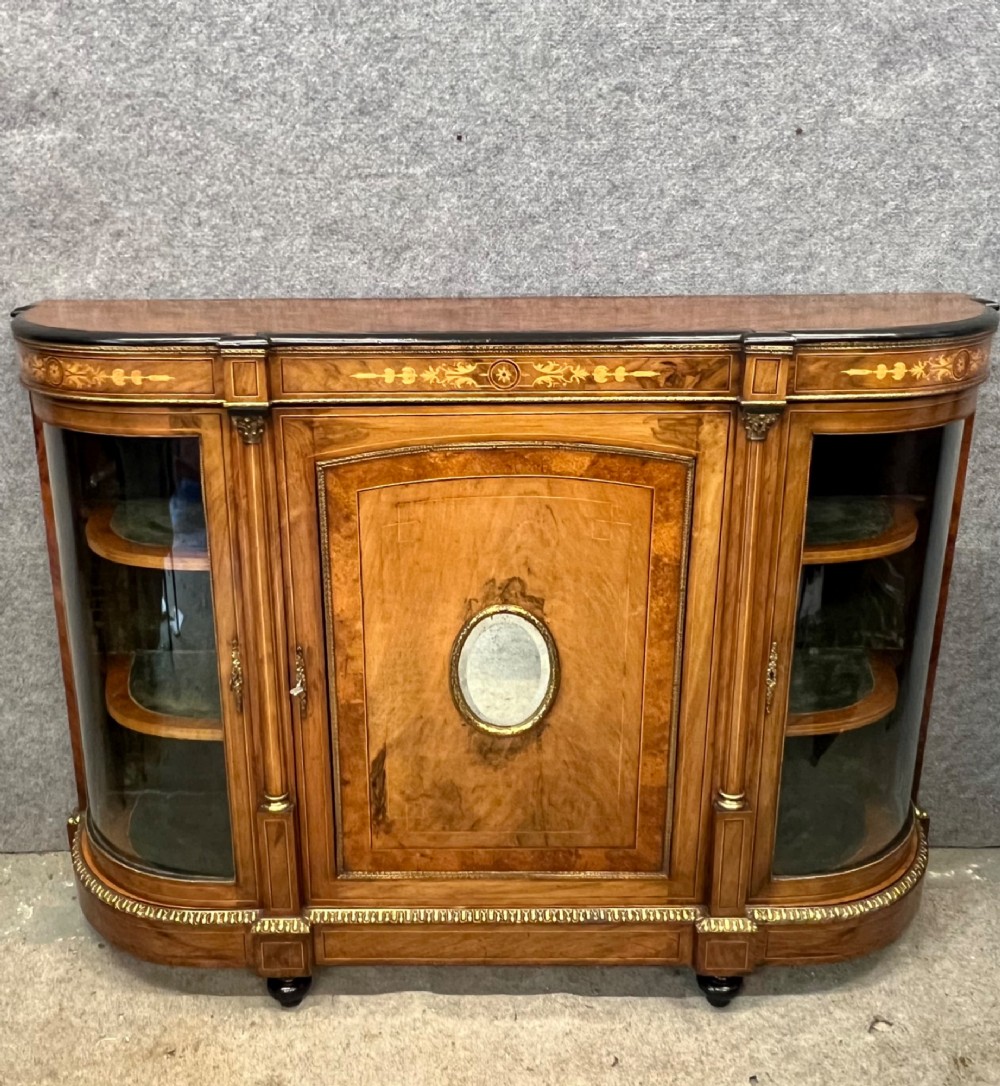 A Victorian credenza crafted from burr walnut with floral marquetry inlay, a central panelled cupboard door, curved glass display bays, and ornate ormolu mounts.