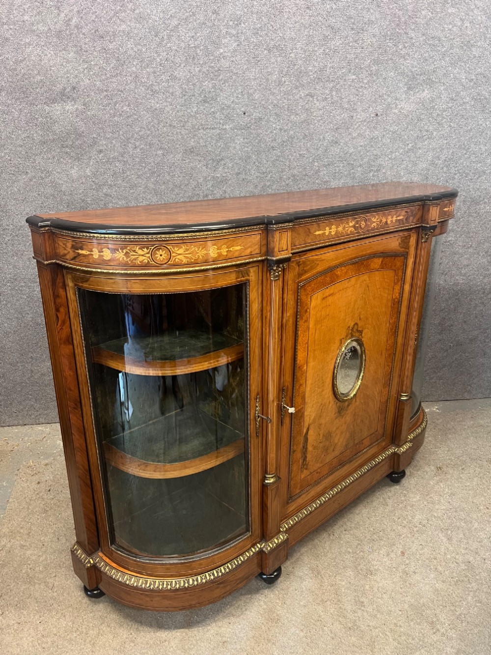 A Victorian credenza crafted from burr walnut with floral marquetry inlay, a central panelled cupboard door, curved glass display bays, and ornate ormolu mounts. View II