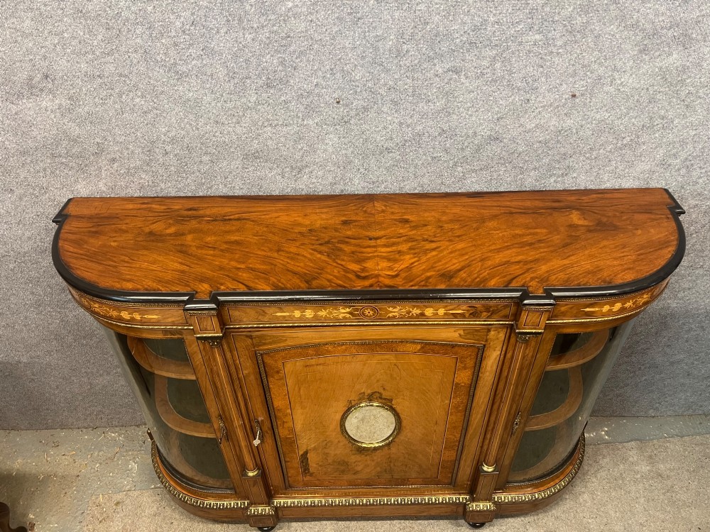 A Victorian credenza crafted from burr walnut with floral marquetry inlay, a central panelled cupboard door, curved glass display bays, and ornate ormolu mounts. View XI