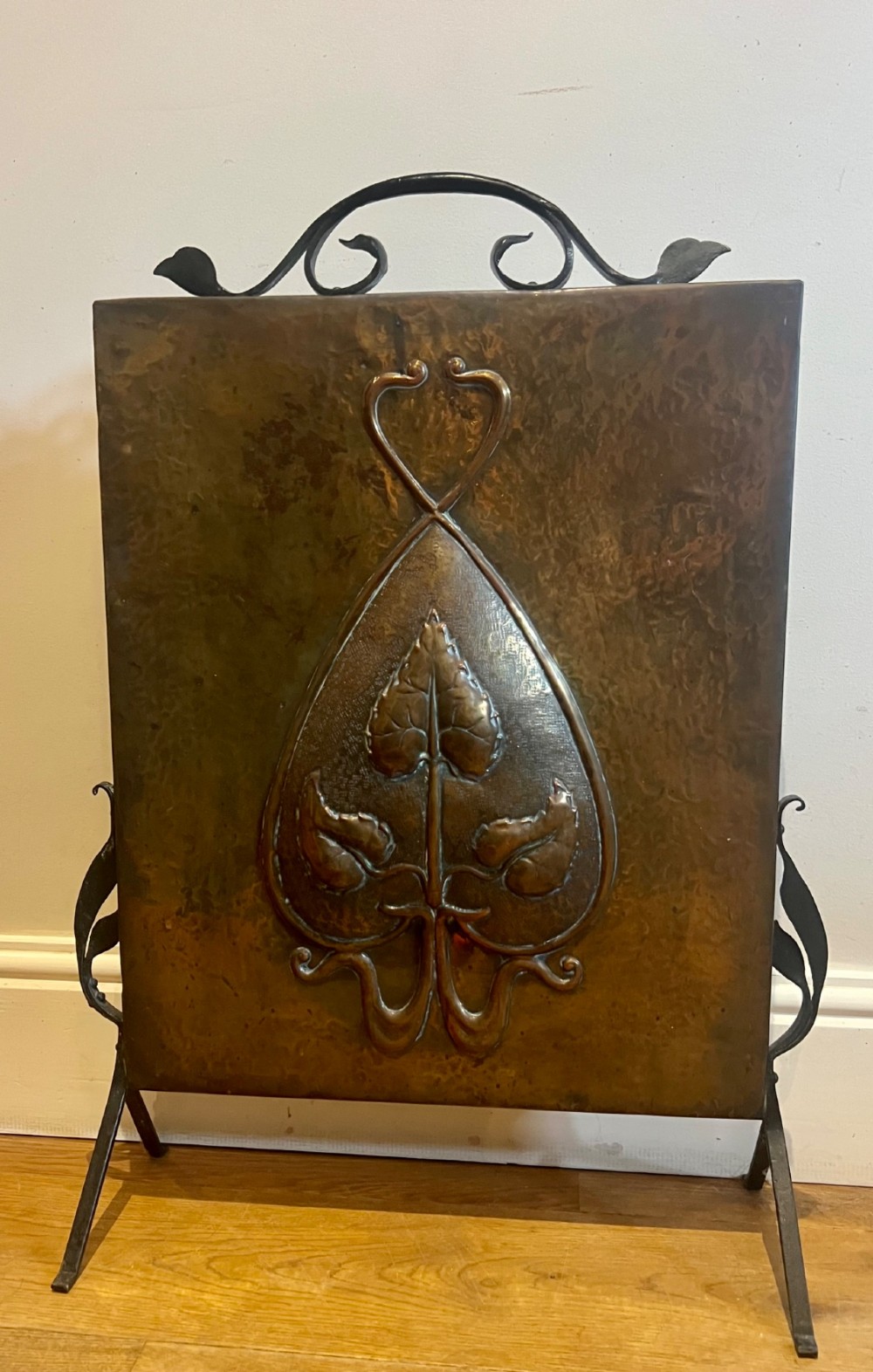 A rectangular hammered copper Arts and Crafts fire screen embossed with stylised botanical leaf motifs, set within a black wrought iron frame with scrolled legs and an arched carrying handle.