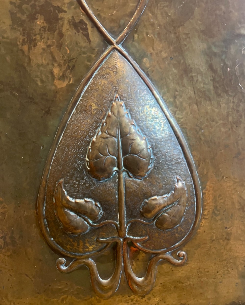 A rectangular hammered copper Arts and Crafts fire screen embossed with stylised botanical leaf motifs, set within a black wrought iron frame with scrolled legs and an arched carrying handle. View II