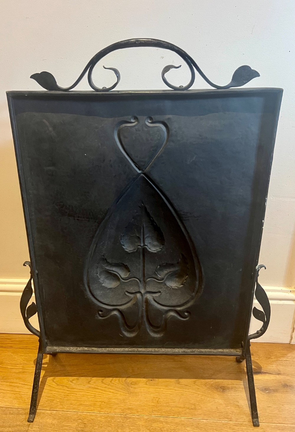 A rectangular hammered copper Arts and Crafts fire screen embossed with stylised botanical leaf motifs, set within a black wrought iron frame with scrolled legs and an arched carrying handle. View V
