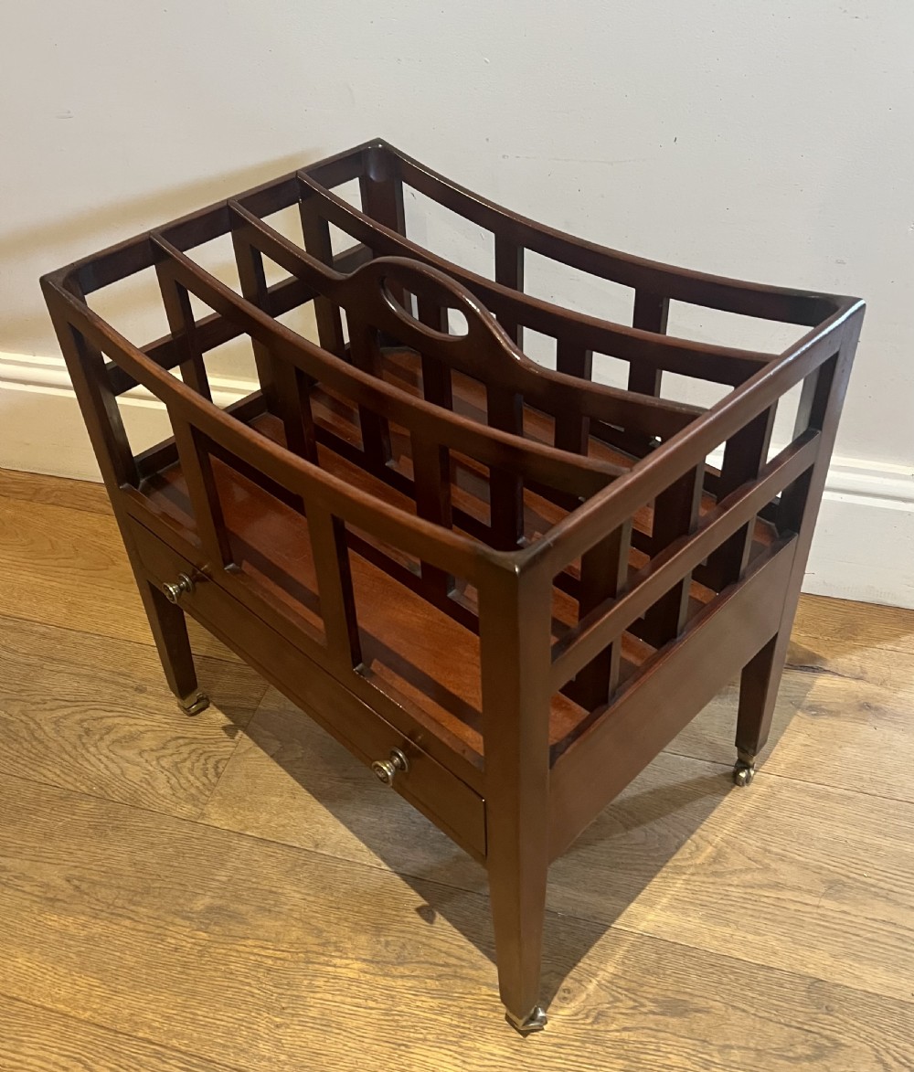 An Edwardian mahogany canterbury with four slatted compartments, a central cut-out handle, a single base drawer with brass knobs, and tapered legs on brass castors.